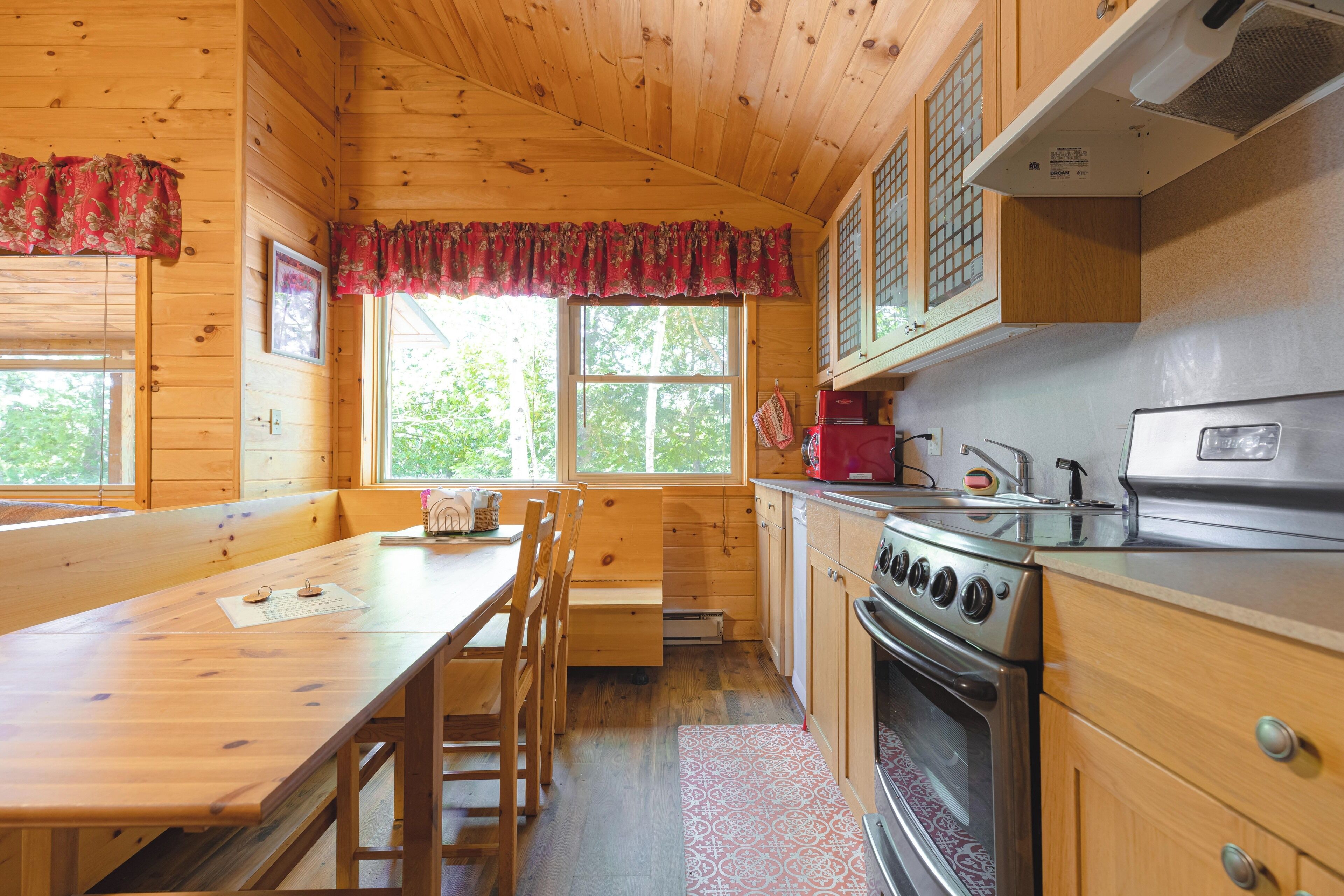 A wooden cabin kitchen with a wooden table, chairs, a stove, and a window with a view of trees.