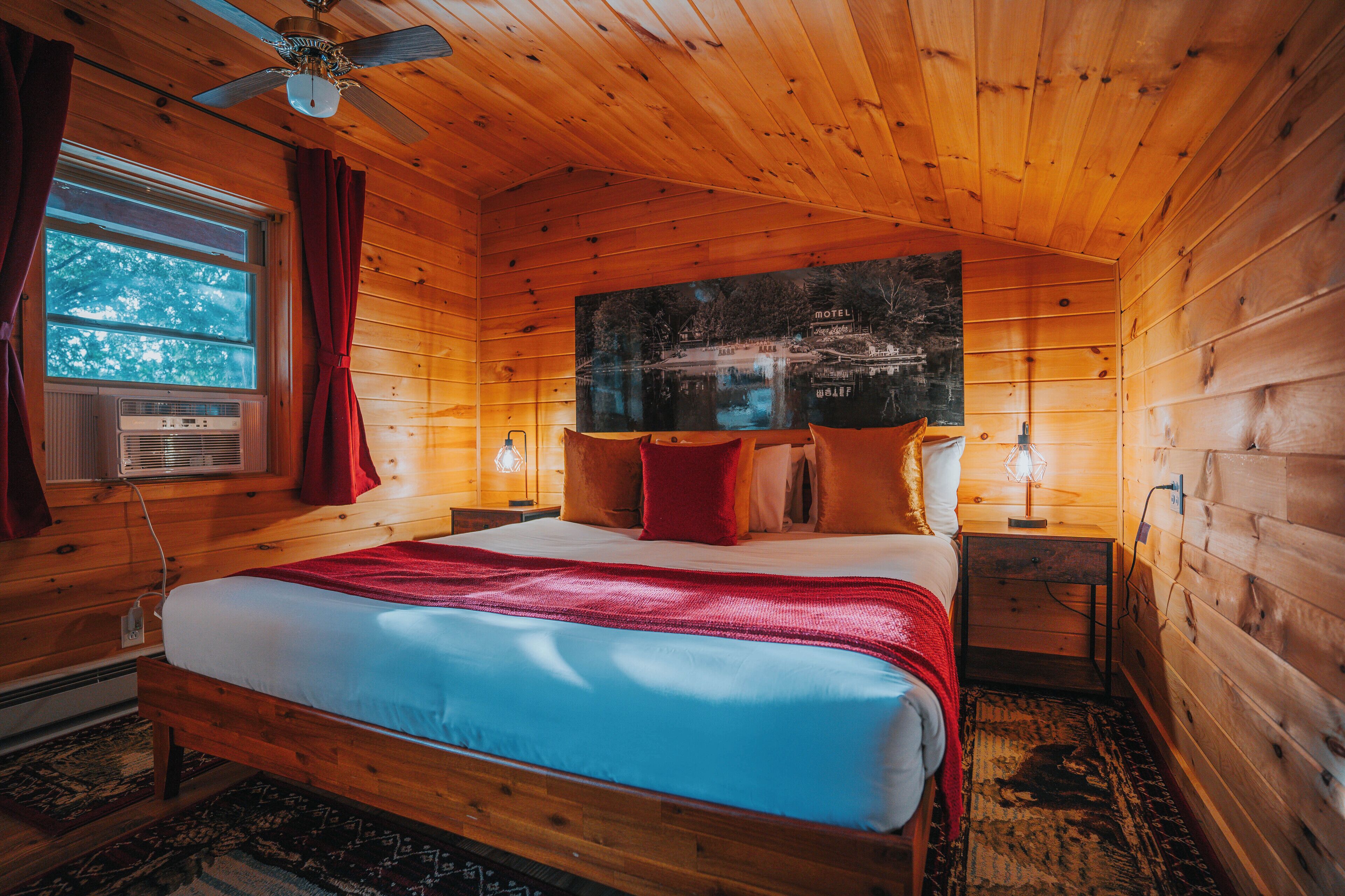 Une chambre rustique en bois, avec un lit, un ventilateur de plafond, une fenêtre avec des rideaux et un tableau accroché au mur.