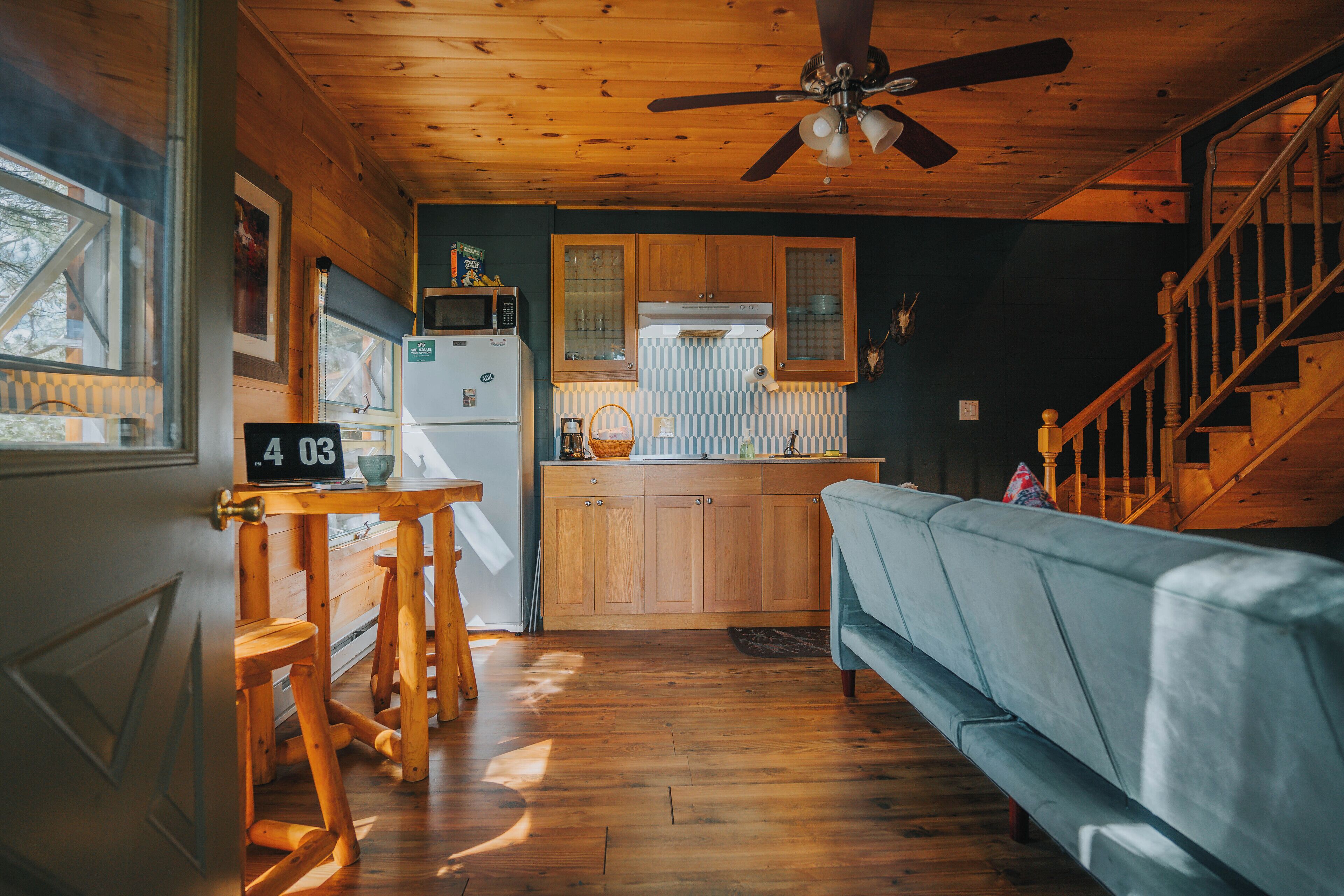 Interior acogedor de una cabaña con techo de madera, ventilador de techo, kitchenette, sofá azul y una escalera.
