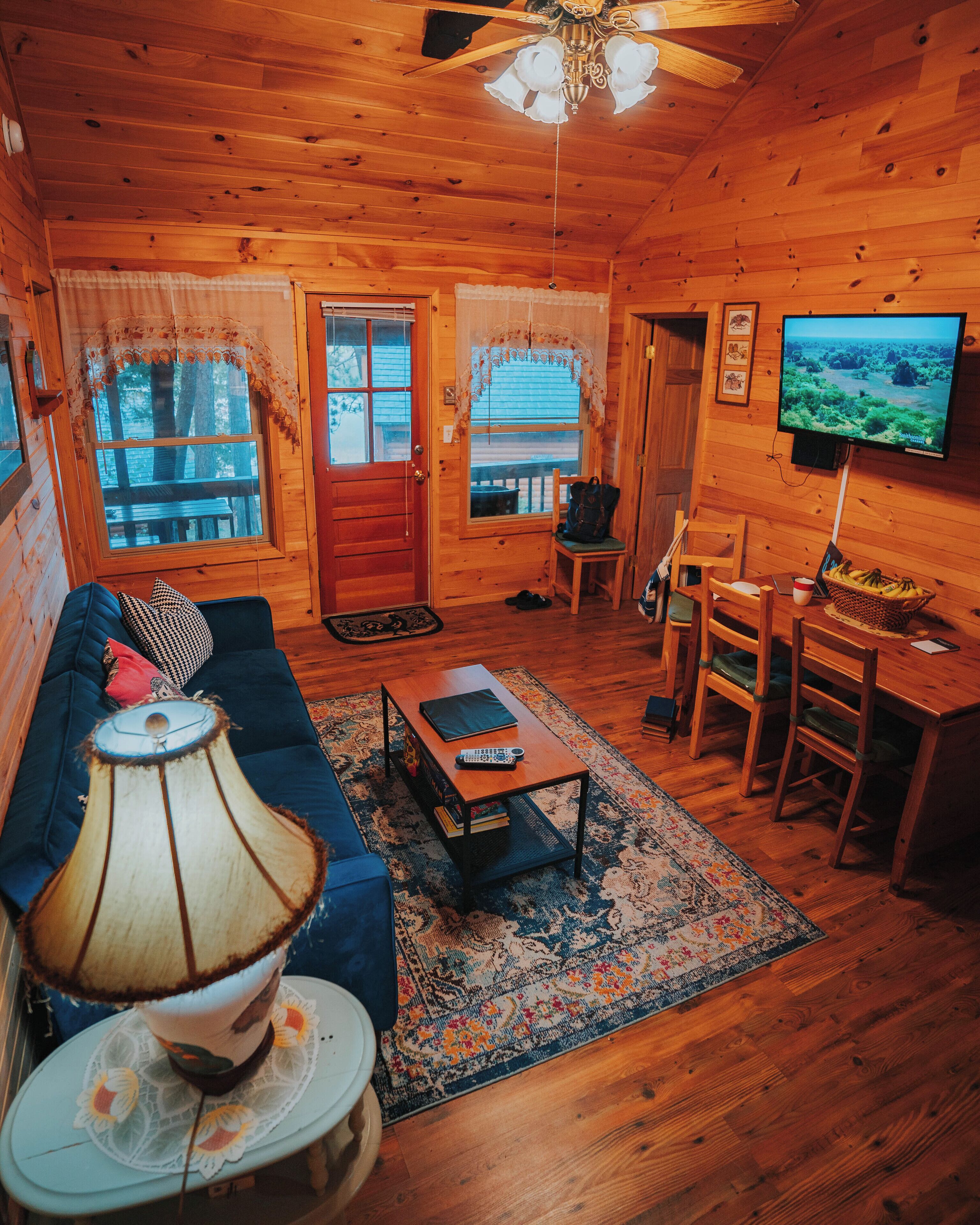A cozy cabin interior with a wooden floor, a sofa, a coffee table, a television, and a dining area.