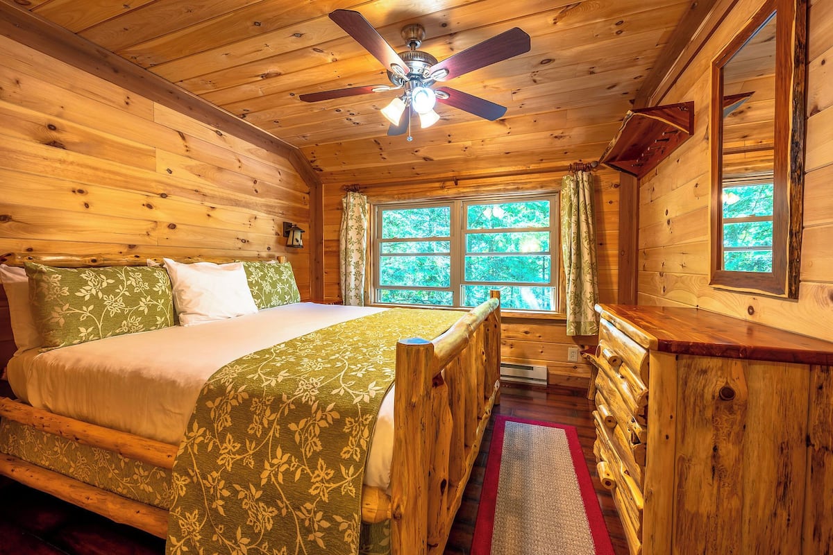 A wooden cabin bedroom with a bed, a ceiling fan, and a window with curtains.