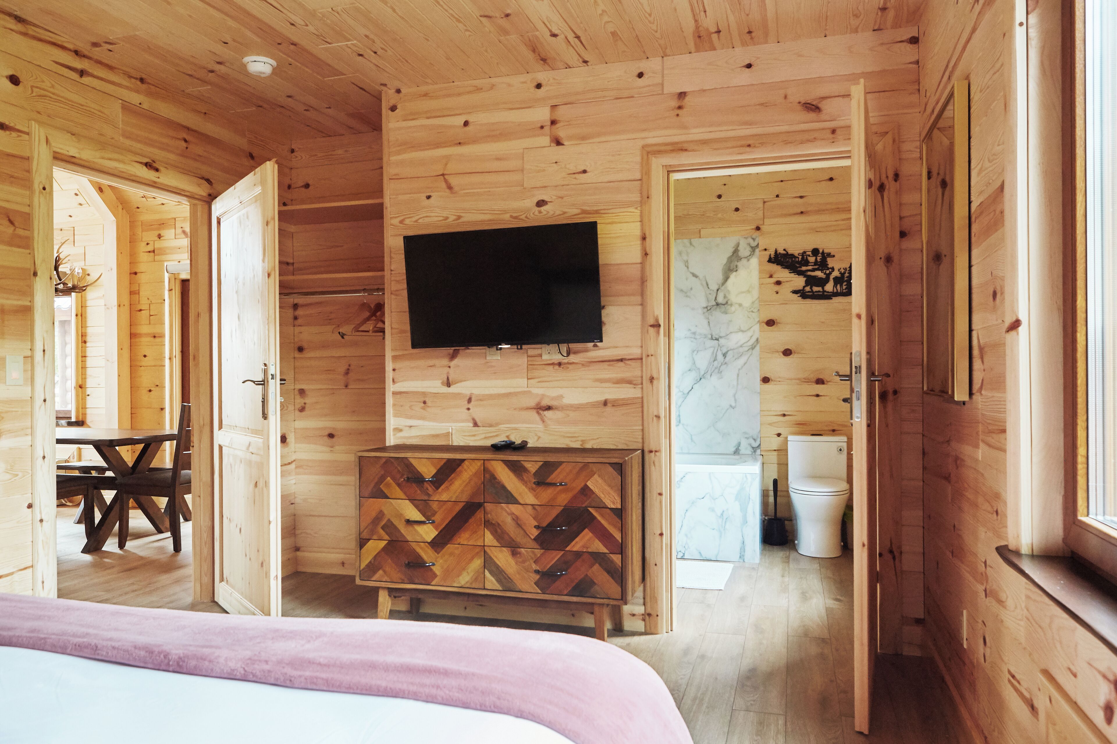 A wooden cabin interior with a bed, a TV mounted on the wall, a herringbone-patterned dresser, a dining table with chairs, and a bathroom with a toilet and sink.