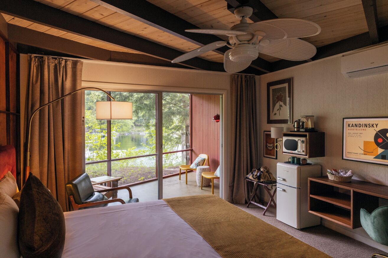 A bedroom with a bed, a ceiling fan, a desk, a chair, a television, and a window with a view of greenery.