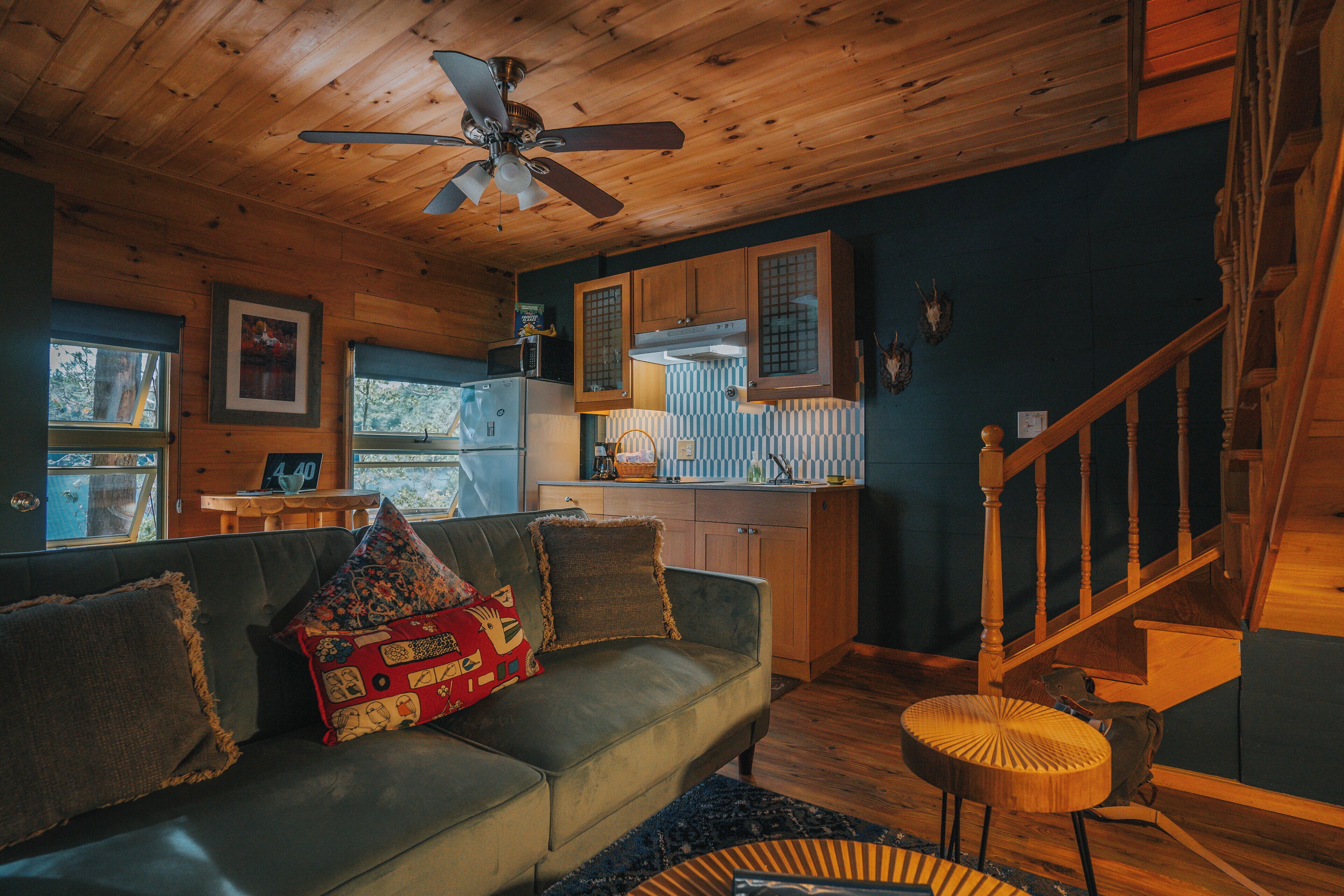 A cozy cabin interior with a wooden ceiling, a sofa with patterned cushions, a kitchen area, and a staircase.