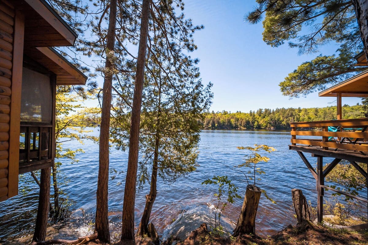 A cabin with a deck overlooking a lake, surrounded by tall trees.