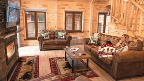 A cozy log cabin interior with a fireplace, two sofas, a coffee table, and a ceiling fan. - Motel Long Lake and Cottages (Long Lake)