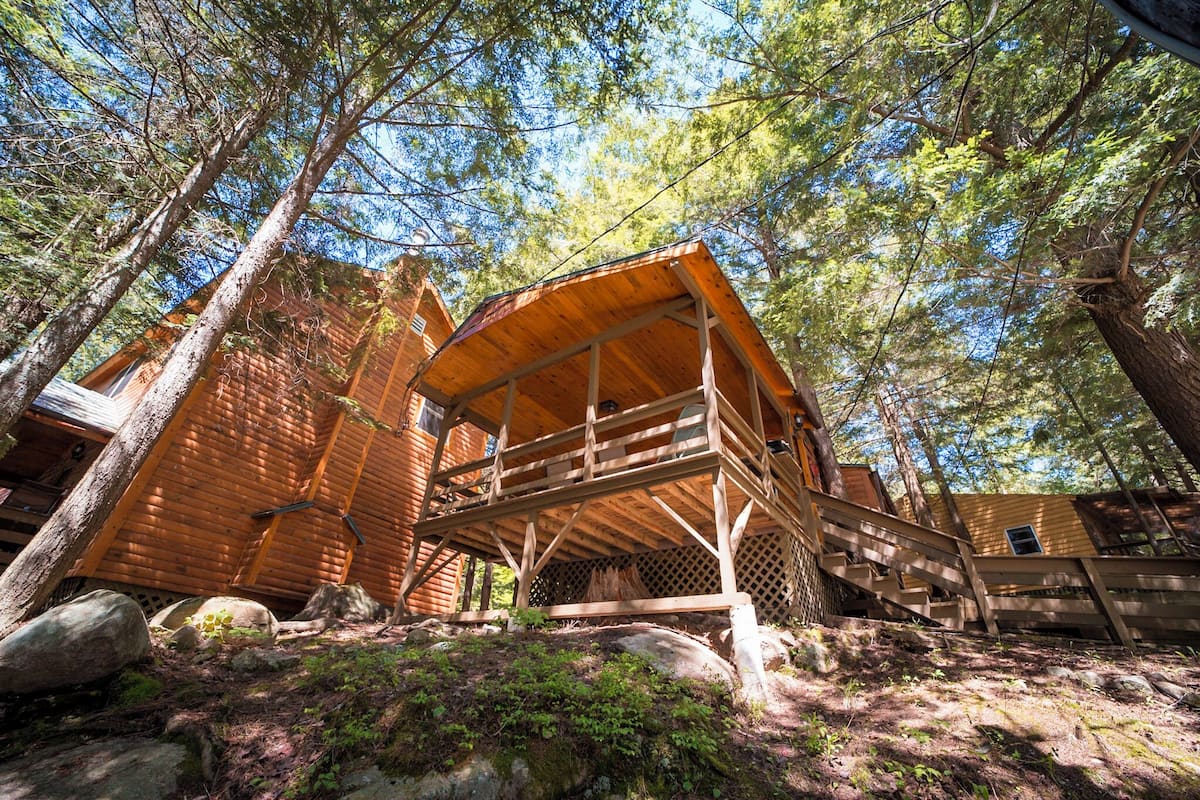 A wooden cabin with a wooden deck and a covered porch, surrounded by trees.