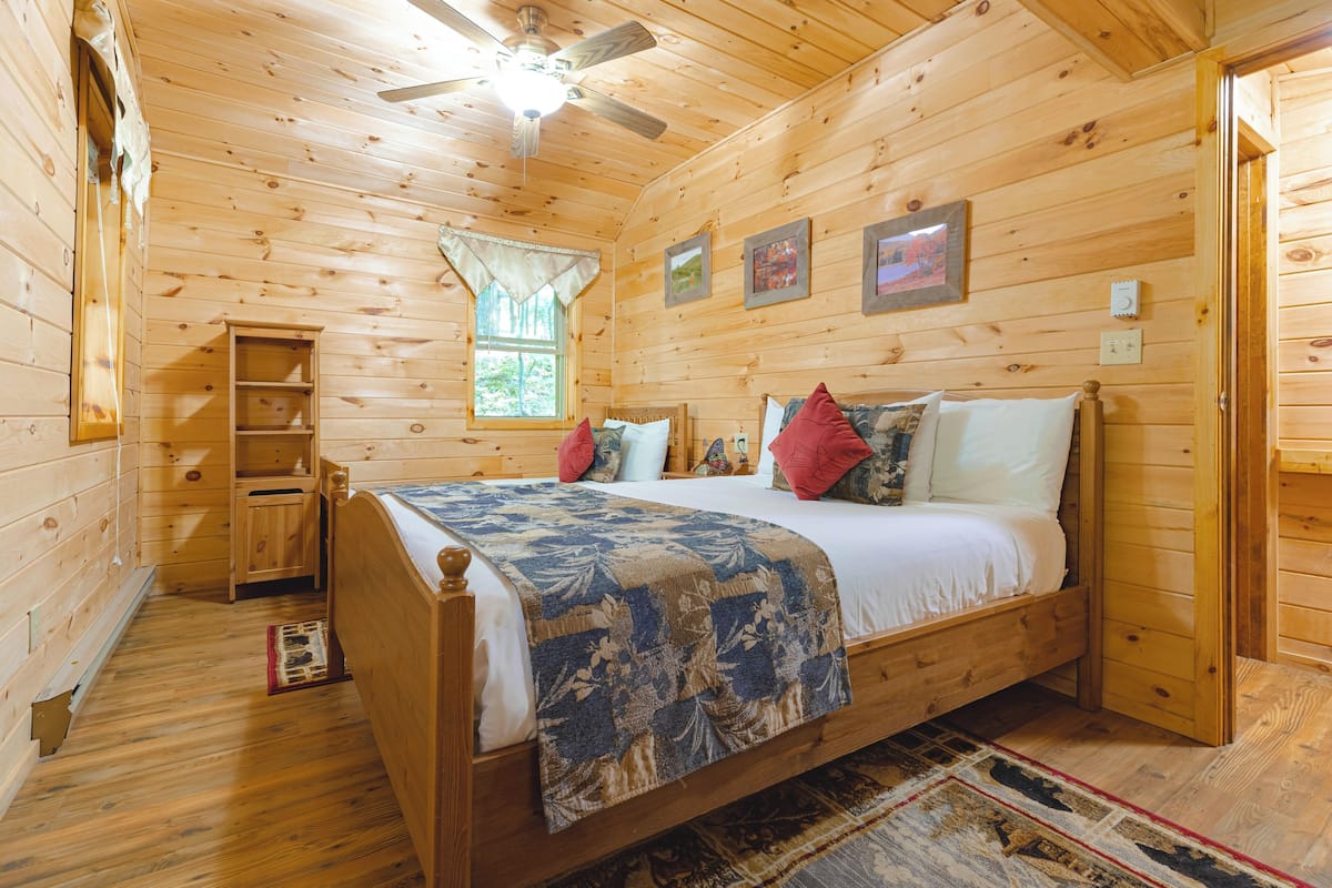 A wooden cabin bedroom with a bed, a ceiling fan, a window with curtains, and a small wooden shelf.