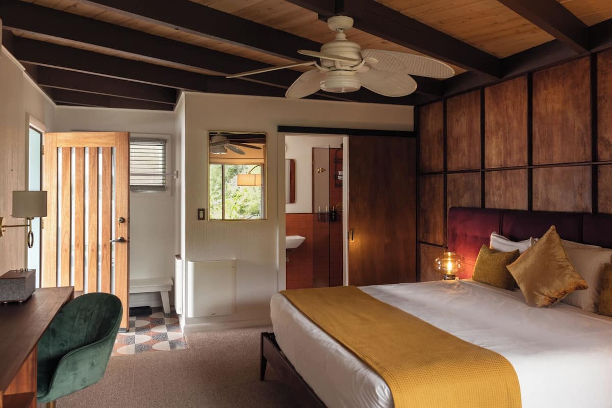 A hotel room with a bed, a desk, a chair, a ceiling fan, and a view of the outside.