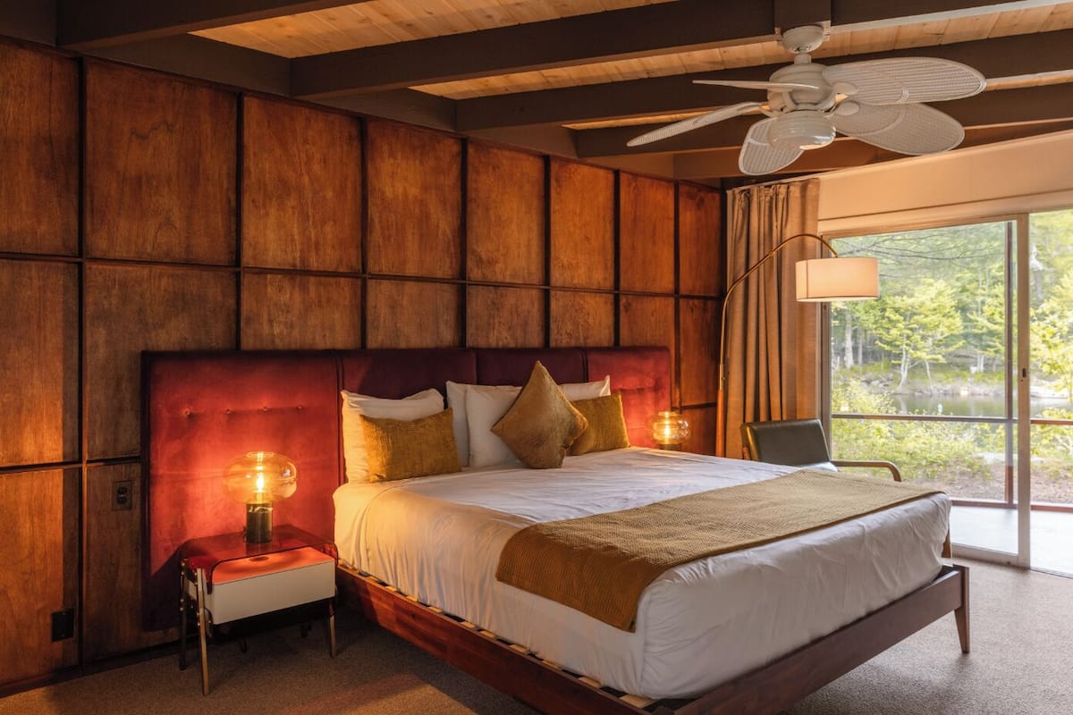 A bedroom with a wooden paneled wall, a bed with pillows, a nightstand, a chair, and a window with a view of trees.