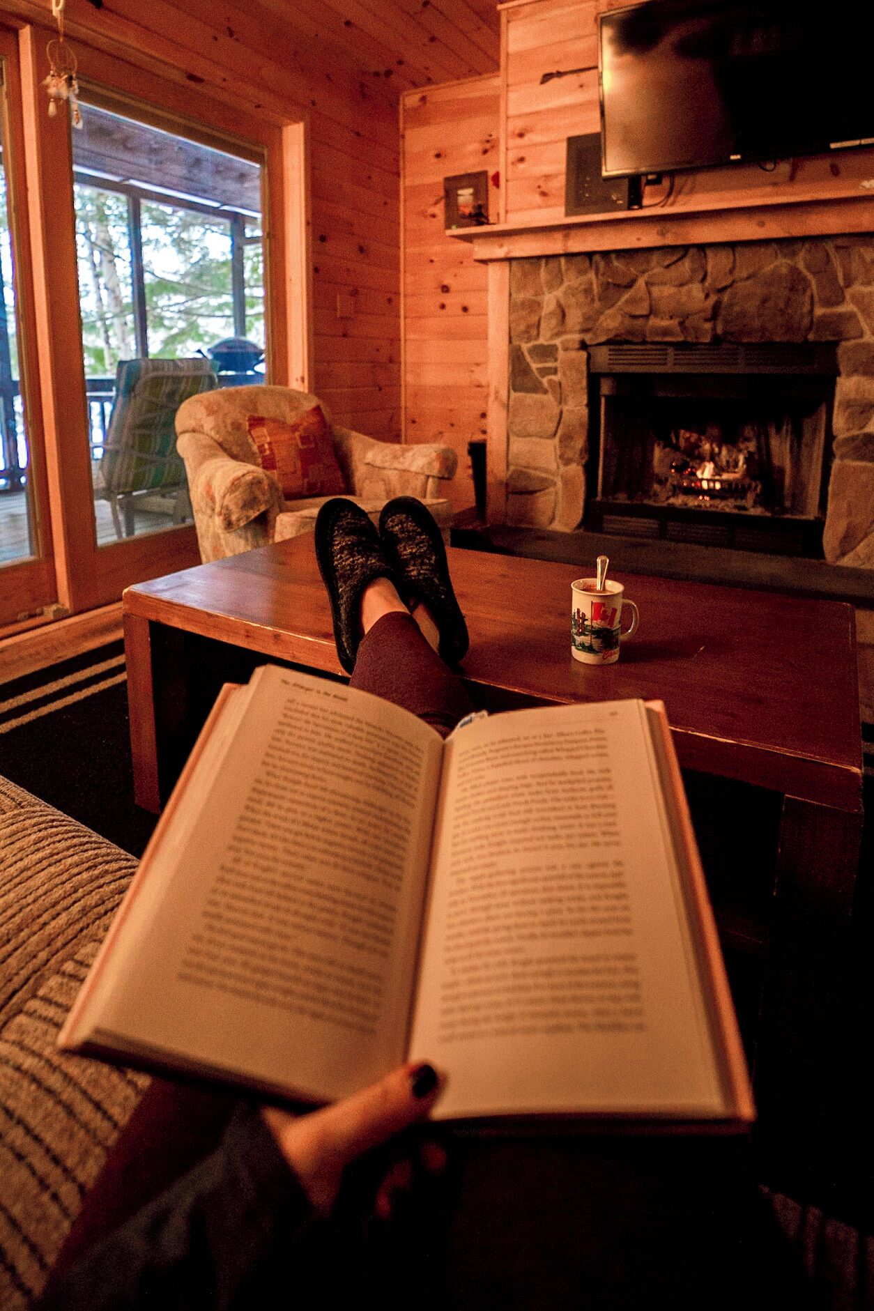Una habitación acogedora con chimenea de piedra, una mesa de madera y una persona leyendo un libro.