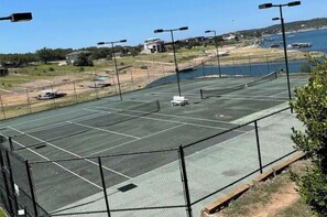 An outdoor tennis court with a net, surrounded by a fence, and overlooking a body of water with buildings in the background. - Great 1 bedroom, Pool/Gym Great Lake Travis Views (Lago Vista)