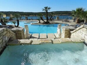 A pool area with stone walls and statues, surrounded by lounge chairs and palm trees, overlooking a body of water. - Great 1 bedroom, Pool/Gym Great Lake Travis Views (Lago Vista)