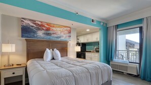 A hotel room with a large bed, a nightstand, a painting, a view of the kitchen, and a balcony. - Beautiful Resort! Cozy, 2 Room Junior Suite! (Myrtle Beach)