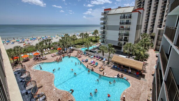 A hotel with a swimming pool surrounded by sun loungers and umbrellas, a beach in the background, and a view of the ocean. - Beautiful Resort! Cozy, 2 Room Junior Suite! (Myrtle Beach)
