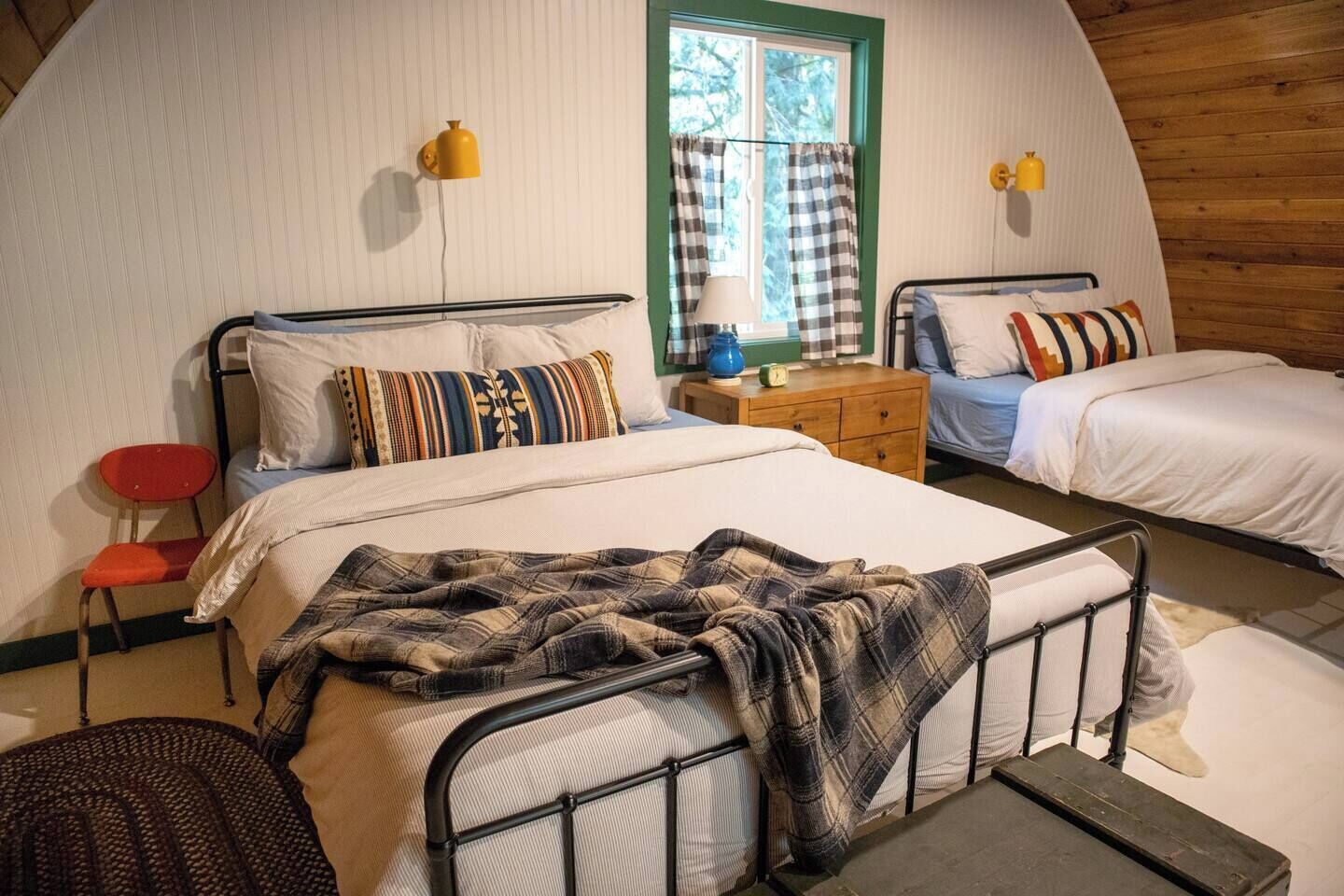 Um quarto aconchegante com duas camas, uma cadeira vermelha, um abajur e uma janela com cortinas.