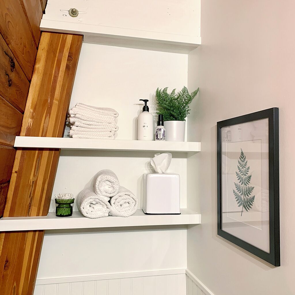 A bathroom with white shelves holding towels, toiletries, and a framed plant.