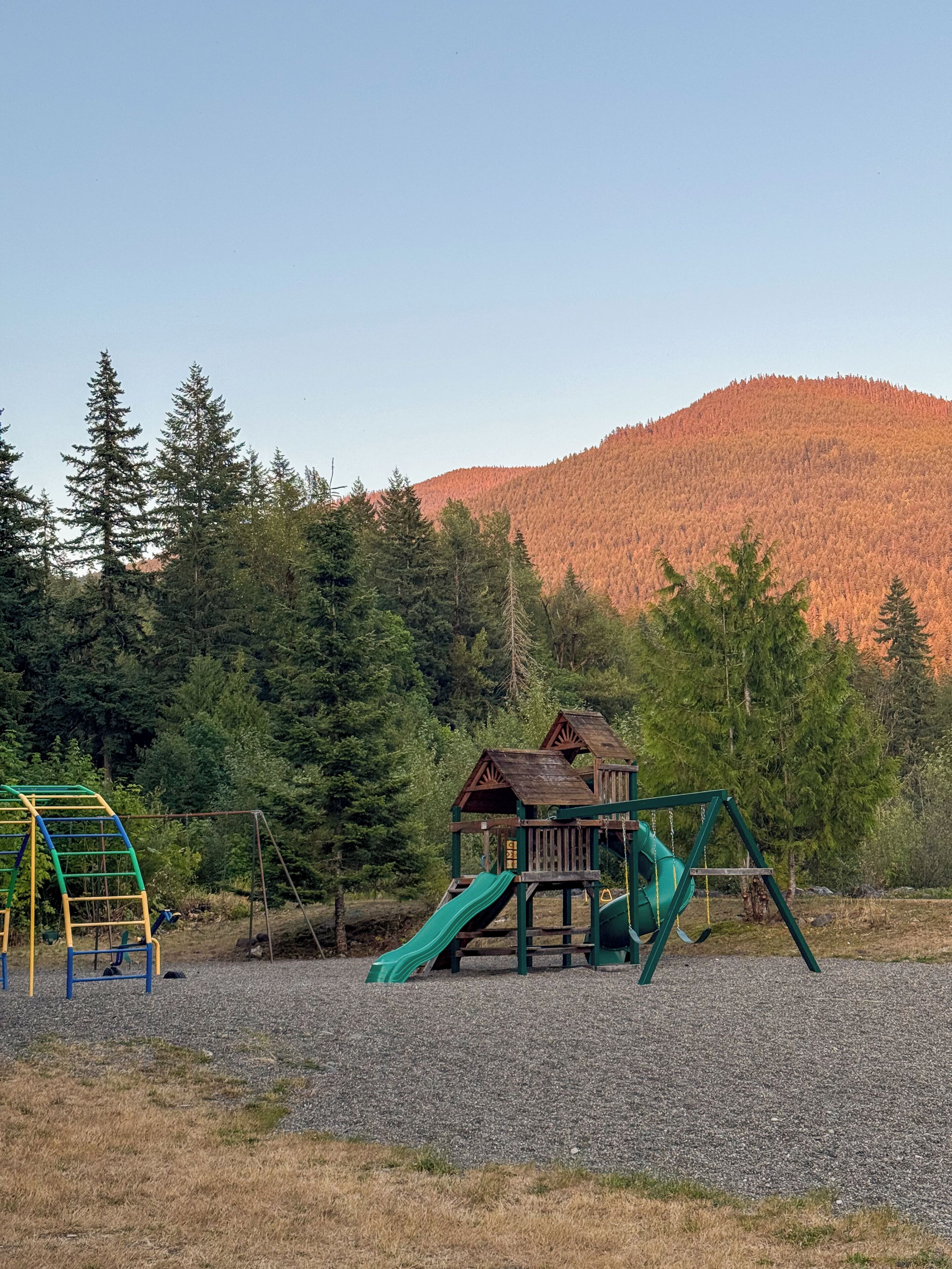Un parque infantil con tobogán y columpios, rodeado de árboles y una colina al fondo.