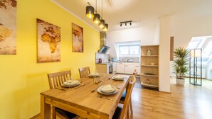 A dining area with a wooden table set for six, a kitchen in the background, and a large window providing natural light.