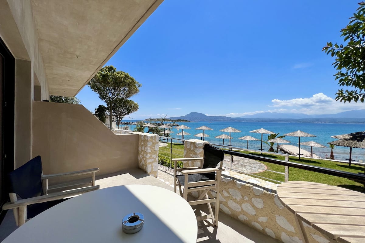 A terrace with a table and chairs, overlooking a pool with umbrellas and a clear view of the sea and distant mountains.