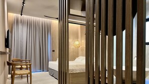 Una habitación de hotel moderna con una cama de madera, una silla, un ventanal con cortinas y una lámpara colgante.