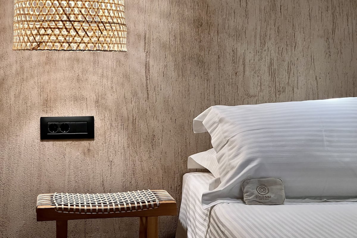 A bedroom with a textured wall, a woven side table, a bed with white linens, a wall-mounted speaker, and a woven light fixture.