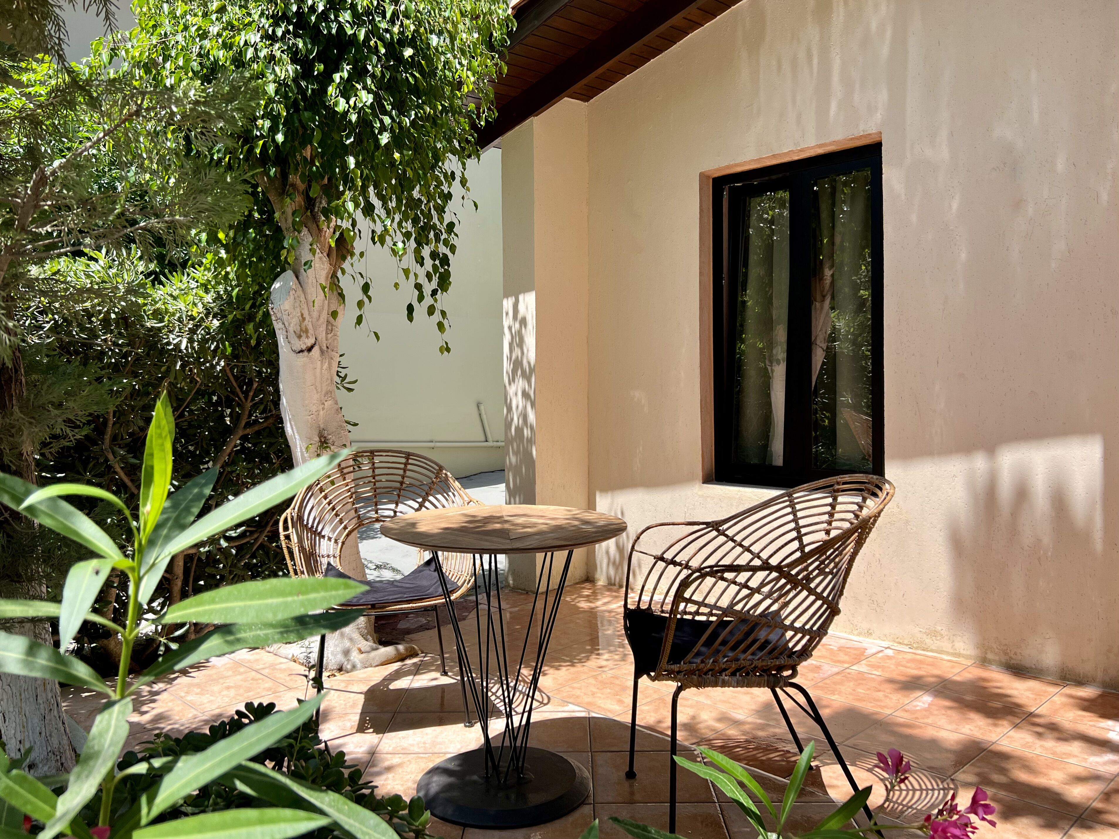 An outdoor patio with a round table and two wicker chairs, surrounded by greenery and a large window.