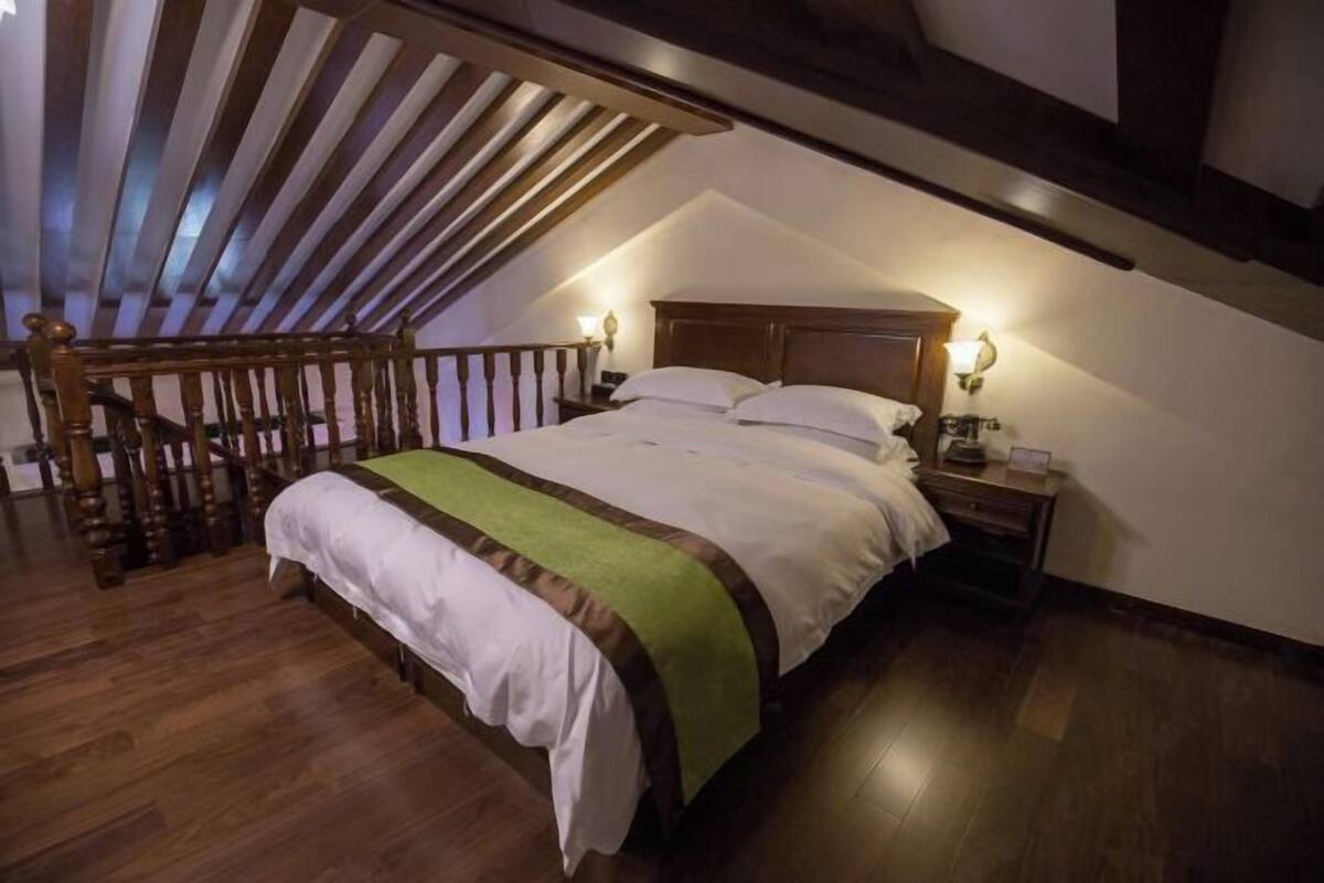 A bedroom with a sloped ceiling, wooden beams, a bed with white bedding and a green throw, two bedside lamps, and a nightstand.