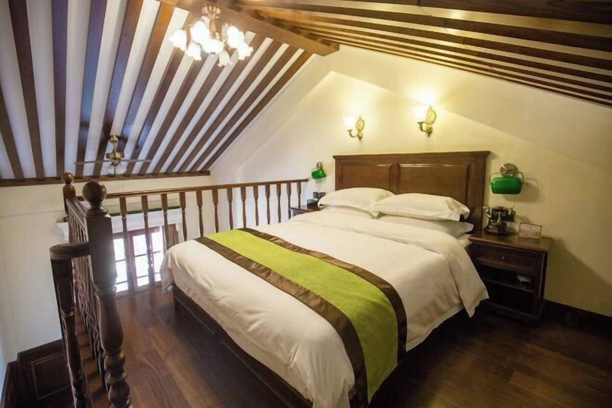 A bedroom with a sloped ceiling, wooden beams, a bed with white bedding, two wall-mounted lamps, and a nightstand with a green lamp.