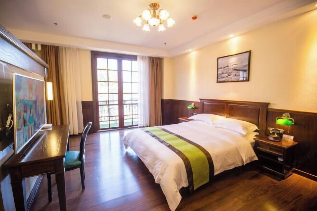 A hotel room with a bed, a desk, a chair, a desk lamp, a nightstand, a painting, and a window with curtains.