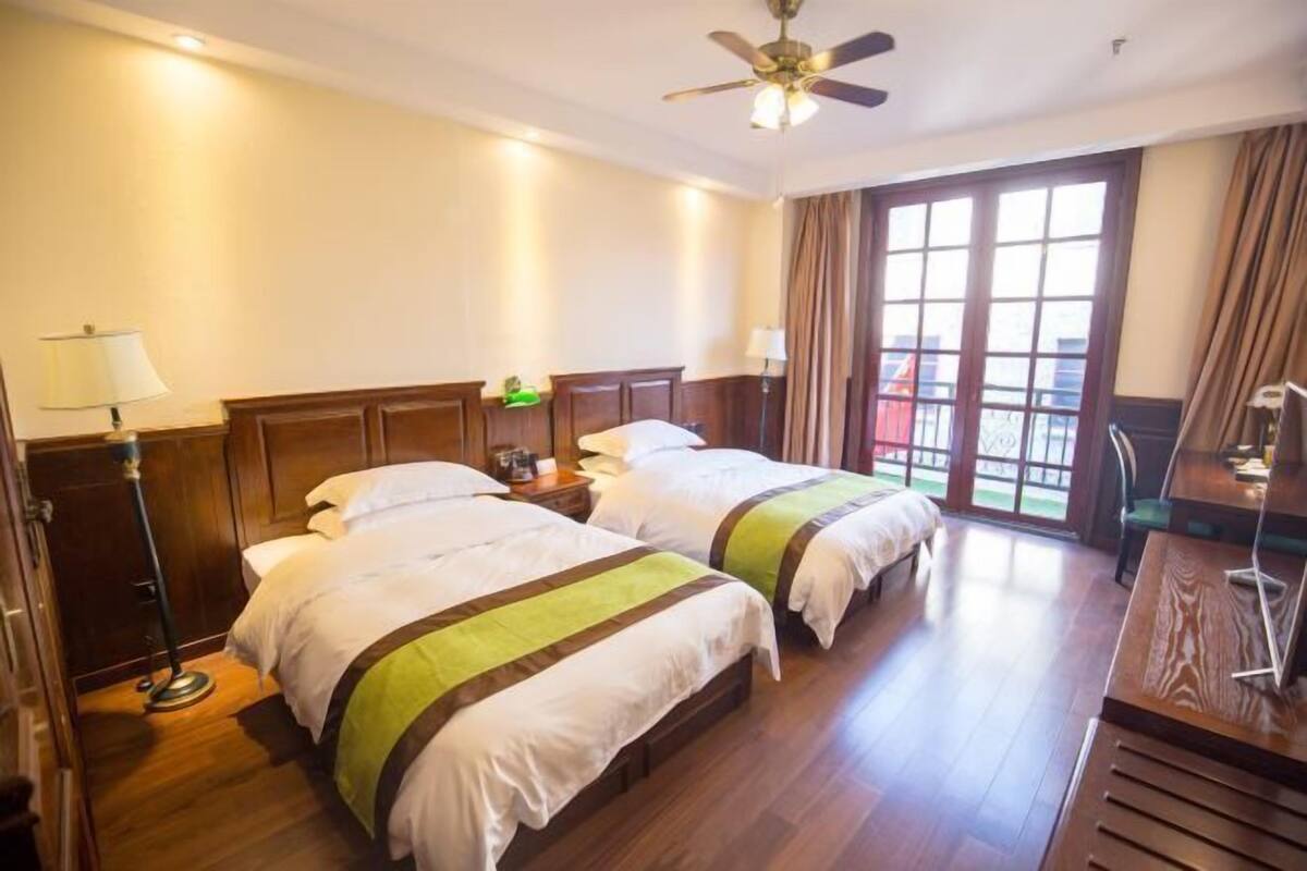 A hotel room with two beds, wooden flooring, a ceiling fan, and a large window with curtains.