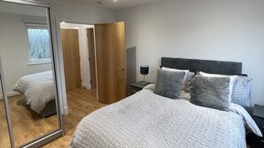 Une chambre avec un grand miroir, un lit recouvert d’une couette blanche à motifs, une armoire en bois et une table de chevet avec une lampe.