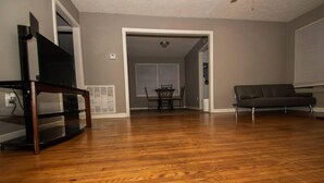 A living room with hardwood flooring, a flat-screen TV on a stand, a gray sofa, and a dining area with a table and chairs. - Home Sweet Home Bungalow (North Little Rock)