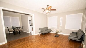 A spacious living room with hardwood flooring, a ceiling fan, a gray sofa, a glass coffee table, and a dining area with a table and chairs. - Home Sweet Home Bungalow (North Little Rock)
