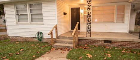 A single-story house with a front porch, a brick foundation, and a small front yard with grass and fallen leaves.