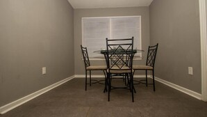 A dining area with a round table and four chairs, a window with blinds, and a plain wall. - Home Sweet Home Bungalow (North Little Rock)