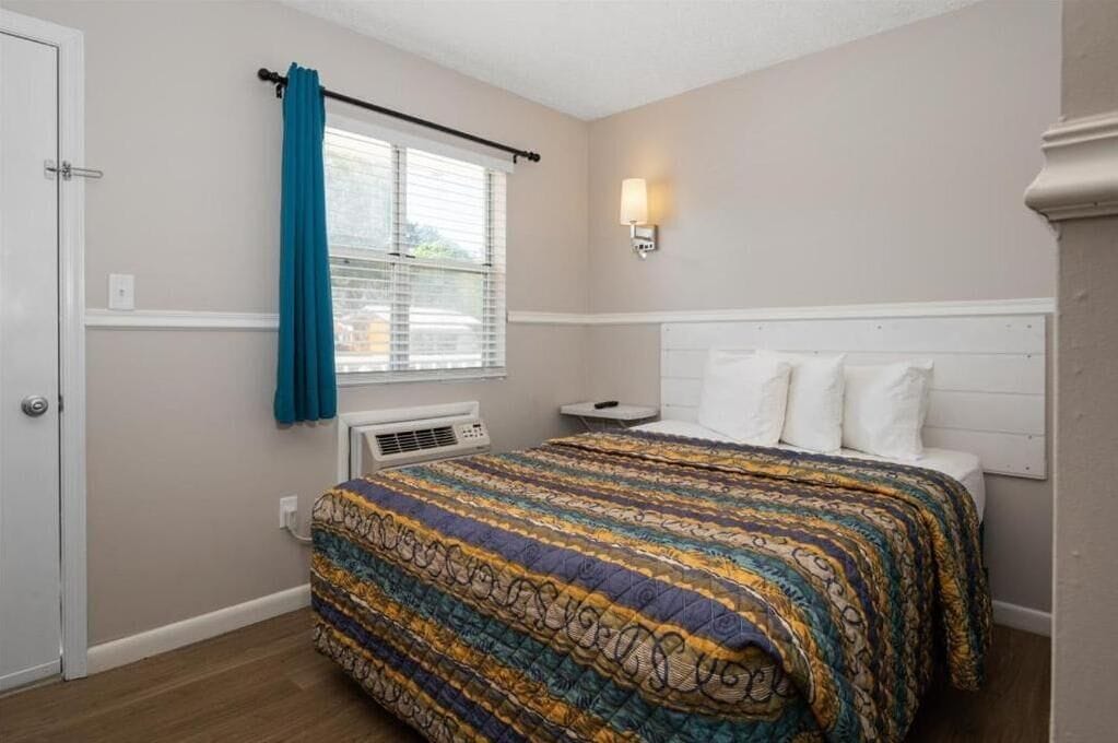 A bedroom with a bed, a window with blinds, a wall-mounted light, and a thermostat.
