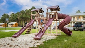 Ein Spielplatz mit Rutschen und Klettergerüsten, umgeben von einer Rasenfläche, mit einem Gebäude im Hintergrund.