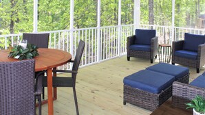 A covered outdoor deck with wicker furniture, a wooden table, and a view of a forest.