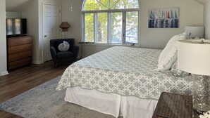 A bedroom with a large bed, a dresser, a chair, a lamp, and a window with a view of trees.