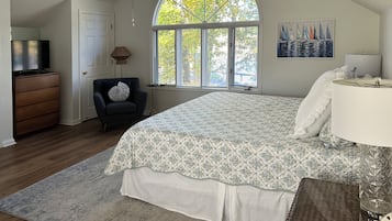 Ein Schlafzimmer mit einem großen Bett, einem Schminktisch, einem Sessel, einer Lampe und einem Fenster mit Blick auf Bäume.