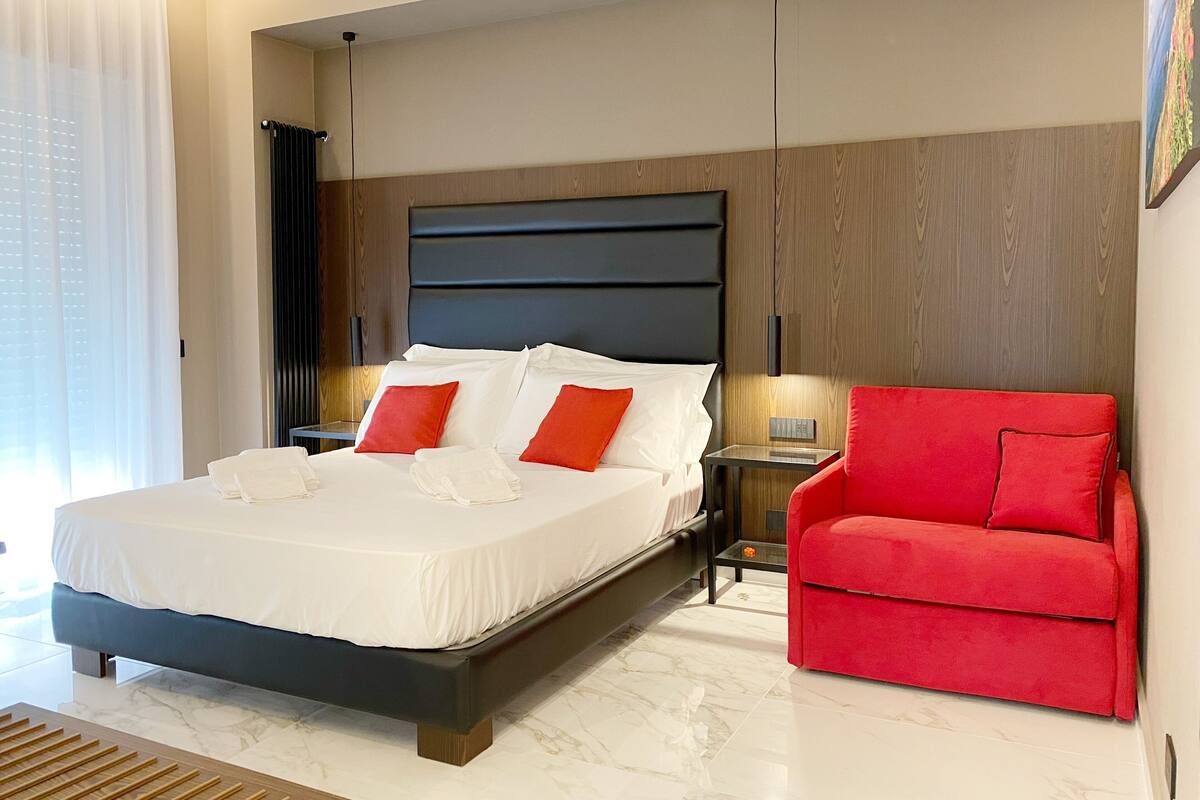 A modern hotel room with a large bed, a red armchair, and a small table.