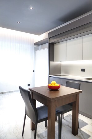 A modern dining area with a wooden table and chairs, a bowl of fruit, and a kitchen in the background. - BorgiaRooms (Milazzo)