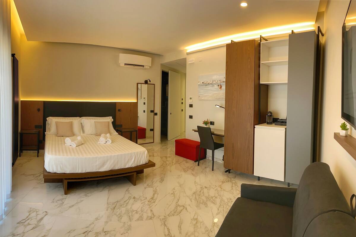 A modern hotel room with a large bed, a desk, and a small kitchenette.