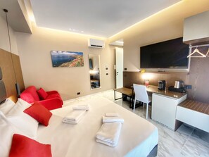 A modern hotel room with a large bed, red pillows, a desk, a television, and a painting of a coastal landscape. - BorgiaRooms (Milazzo)