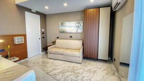 A modern bedroom with a marble floor, a beige sofa, a wooden cabinet, and a painting of a beach scene. - BorgiaRooms (Milazzo)