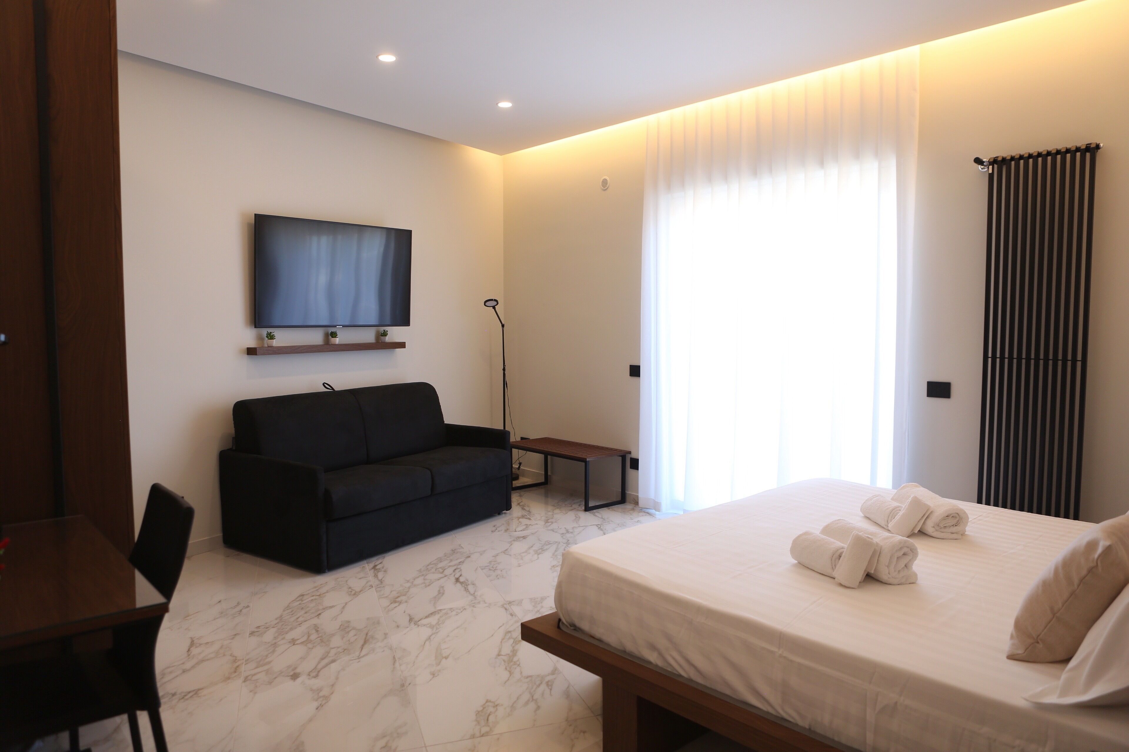 Una camera d'albergo moderna con un letto, un divano nero, una TV a muro e una lampada da terra.