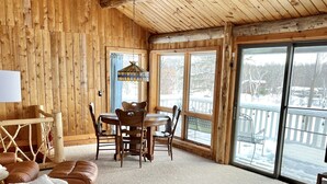 Ein gemütliches Blockhaushaus-Interieur mit Holzbalkendecke, Essbereich, einer Couch mit Lederkissen und großen Fenstern mit Blick auf eine verschneite Terrasse.