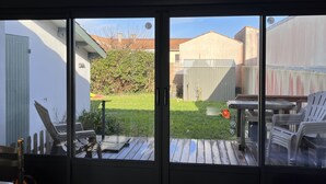 A view through a sliding glass door of a backyard with a wooden deck, a white chair, and a small table.