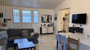 A living room with a sofa, coffee table, and ottoman, a dining area with a table and chairs, a TV mounted on the wall, and a kitchen area with cabinets and a refrigerator.